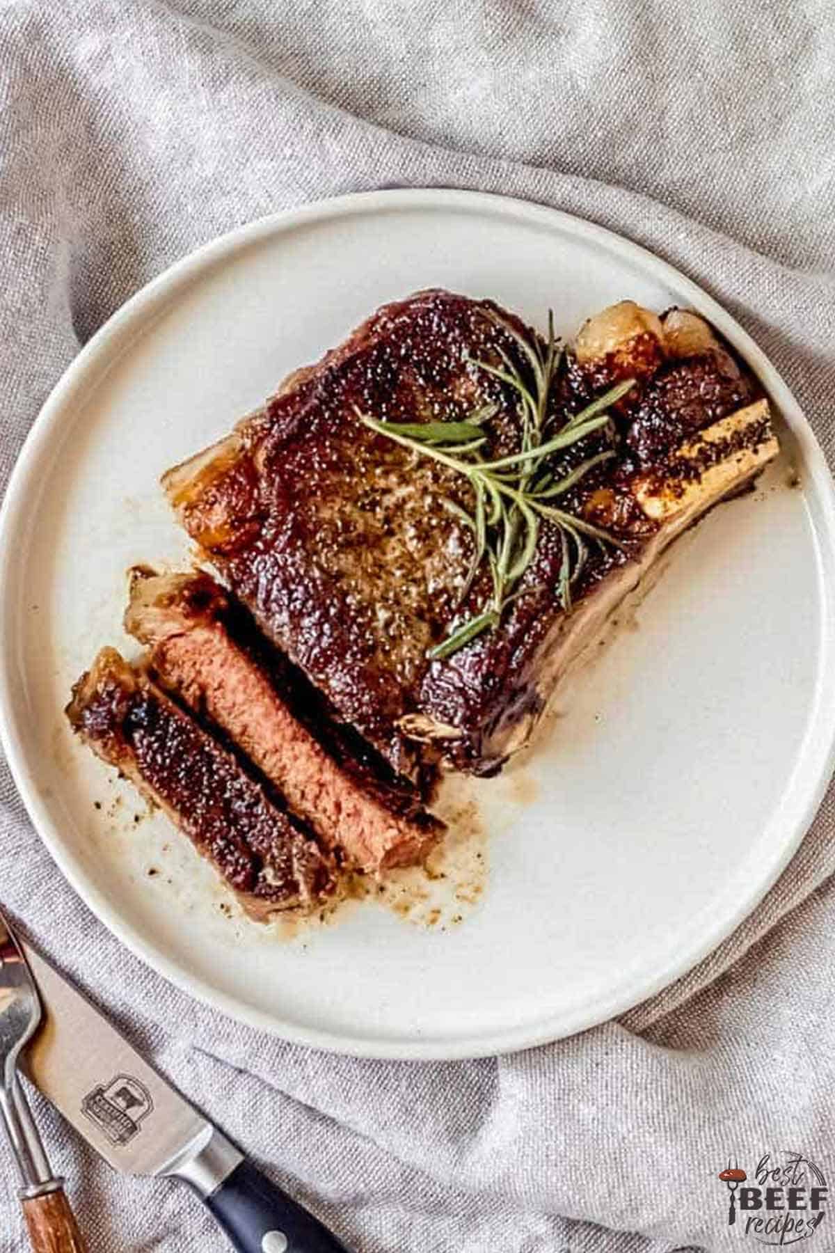 Reverse sear ribeye on a white plate, sliced with sprigs of fresh rosemary on top
