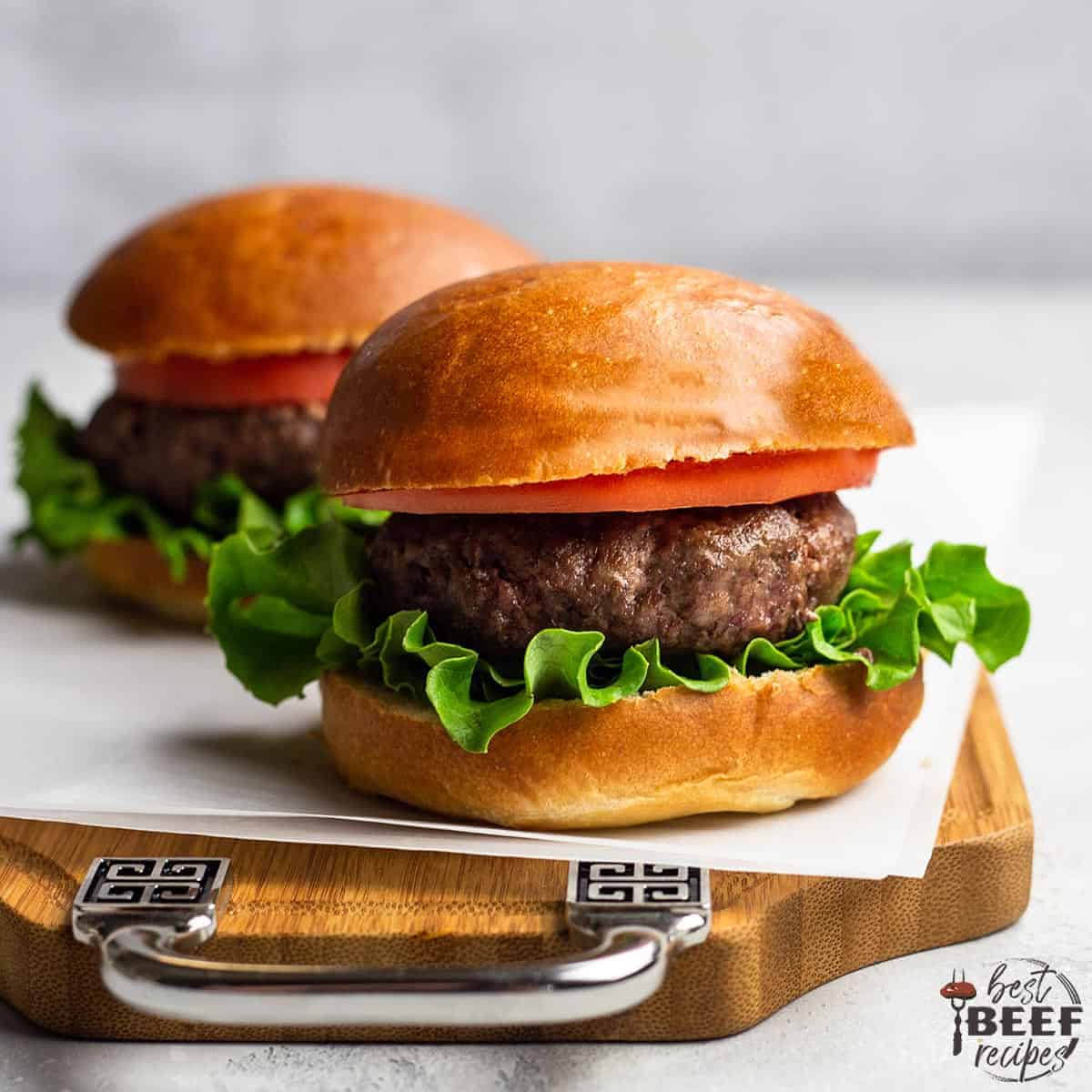 Two burgers made using air fryer burger recipe on golden-brown buns with lettuce and tomato on a piece of parchment paper on a wooden serving board