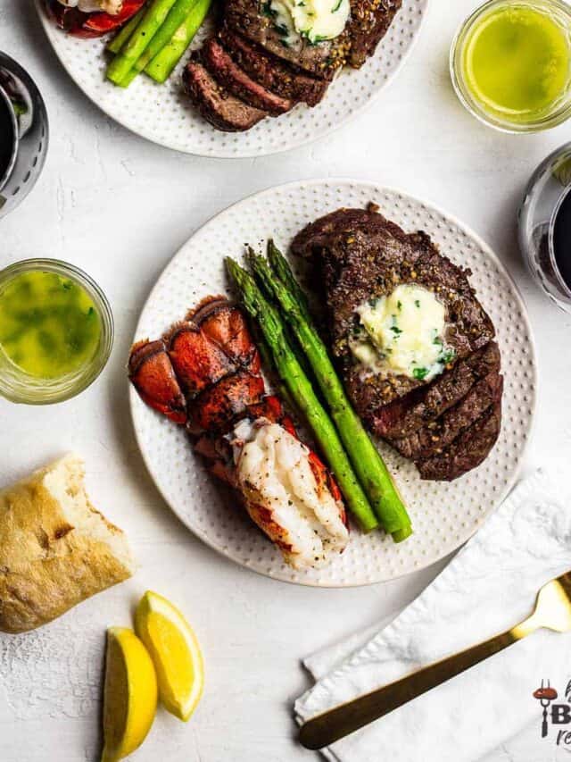 Two plates of air fried steak topped with garlic butter next to asparagus spears and lobster tails