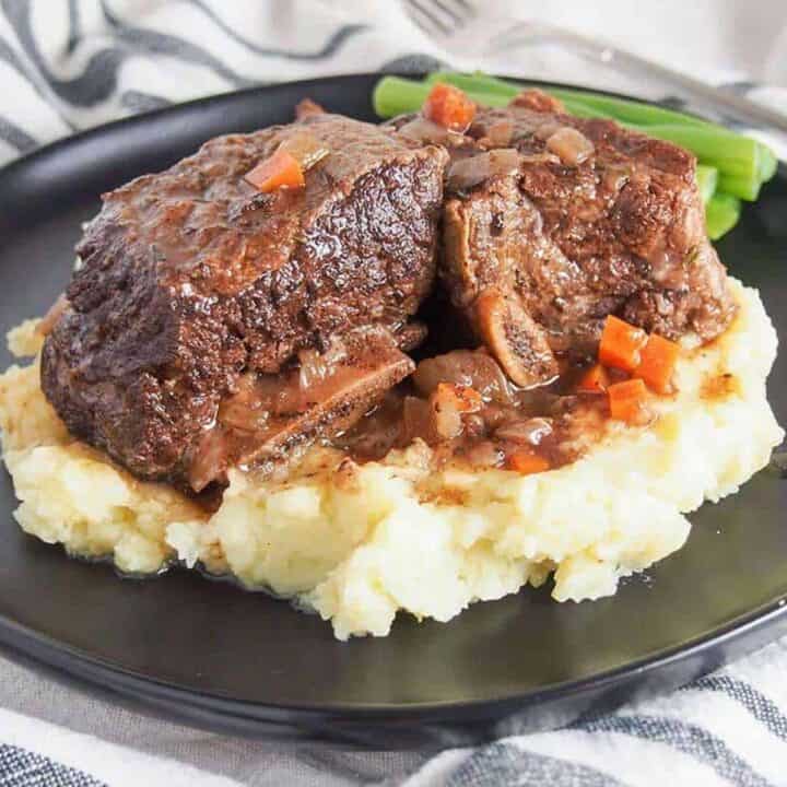 Slow cooker beef short ribs on a black plate over mashed potatoes with green beans on the side