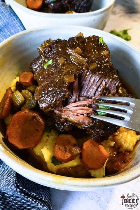 Taking a forkful of beef ribs in a white bowl with vegetables and mashed potatoes