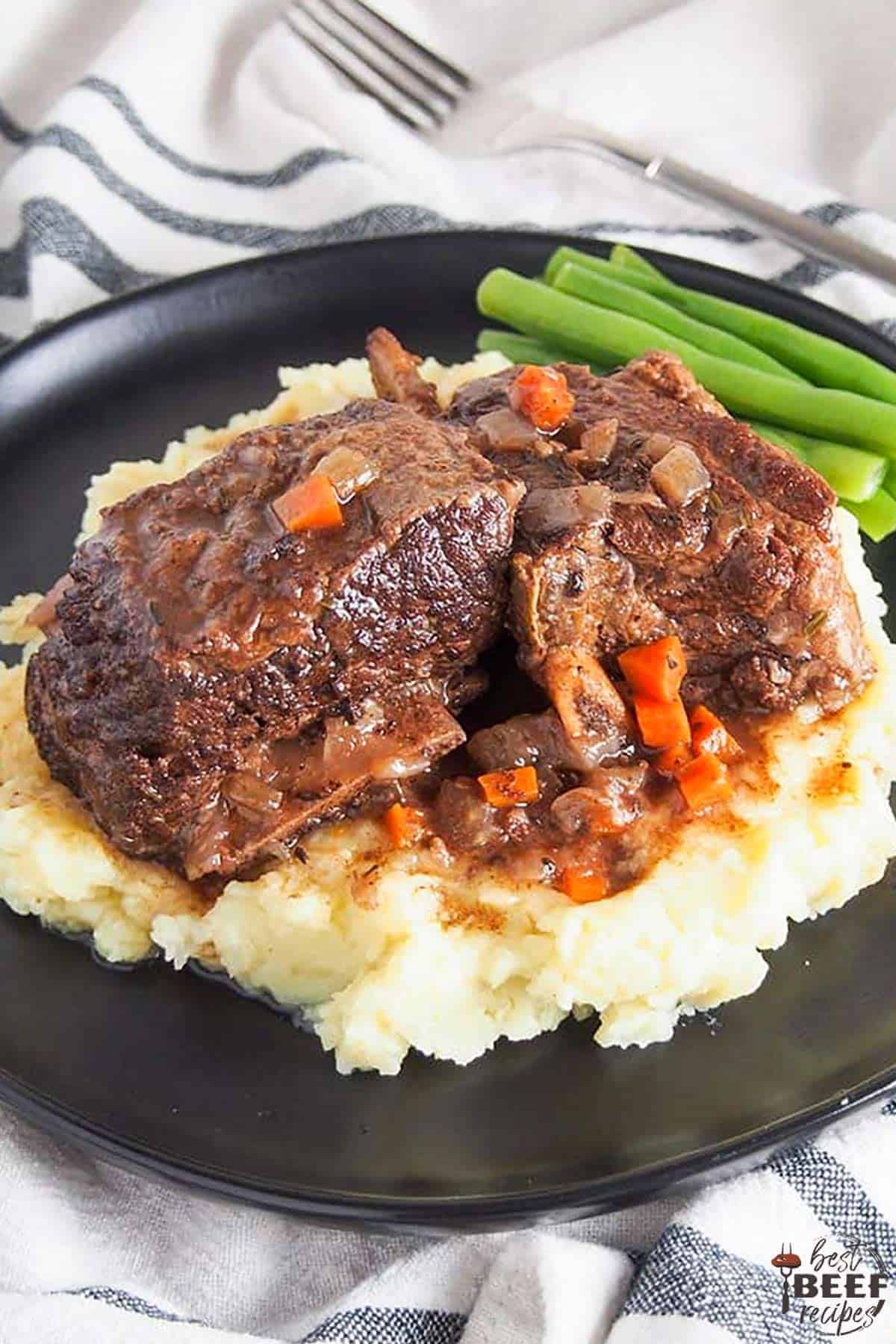 Slow cooker beef ribs served over a bed of mashed potatoes on a black plate with a side of green beans and a decorative white dish towel underneath