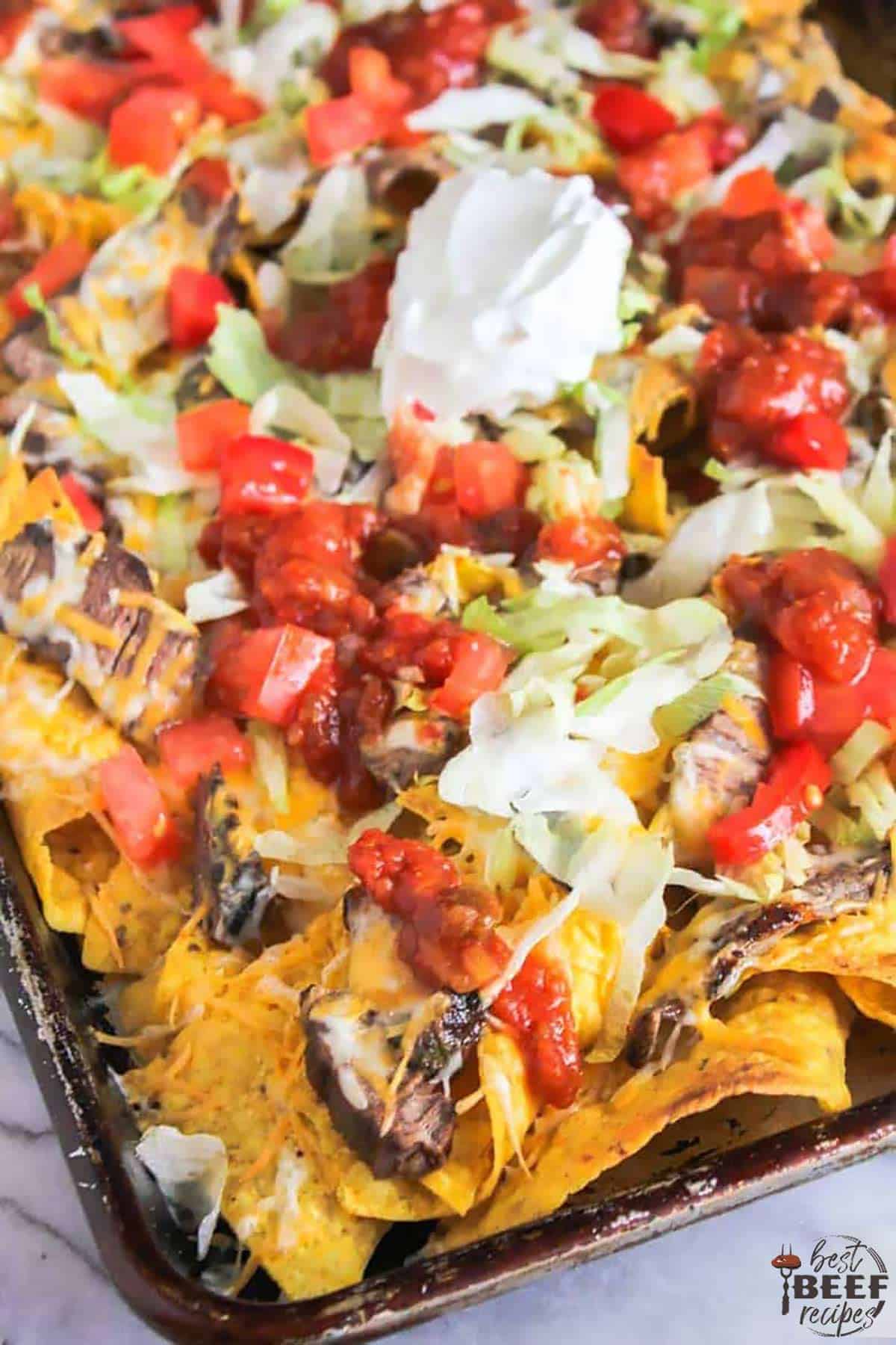 A close up shot of steak nachos with sour cream, chopped tomatoes, and lettuce
