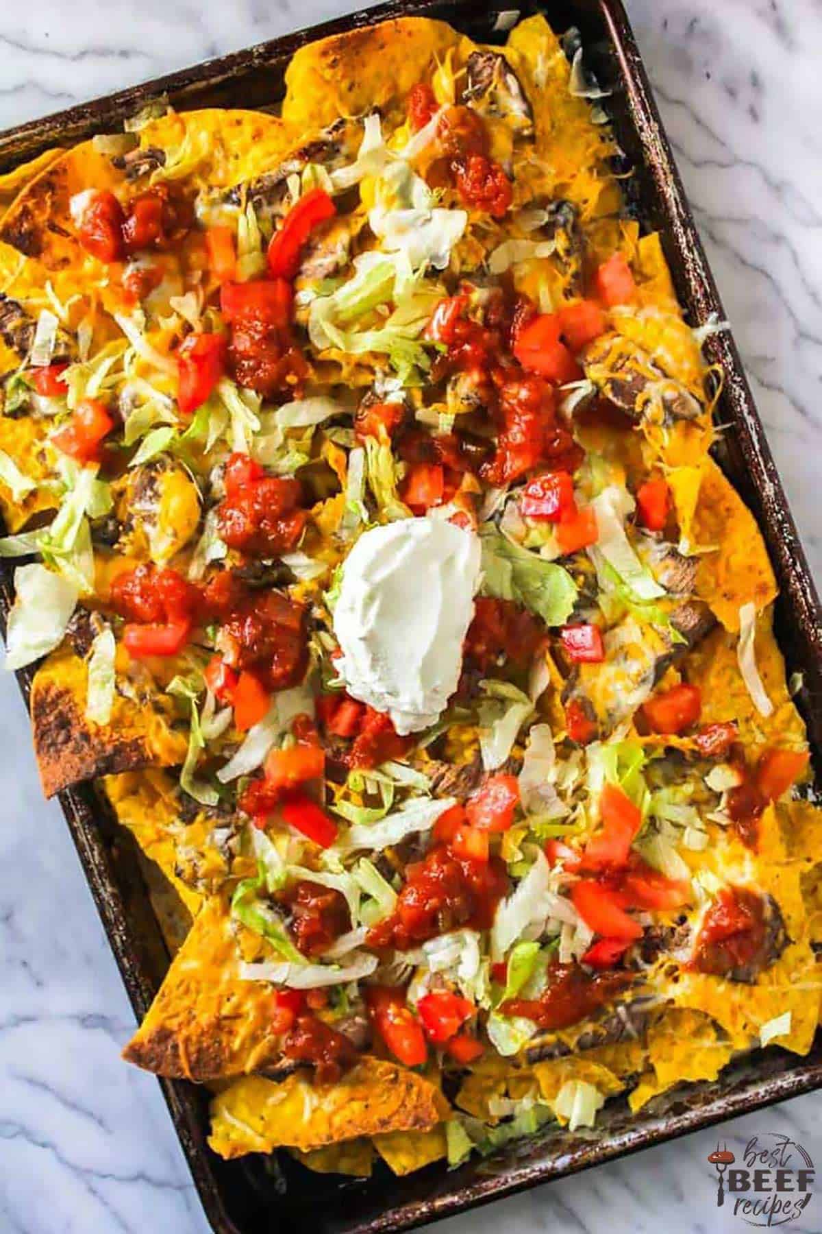 An overhead shot of steak nachos on a sheet pan with a dollop of sour cream on top