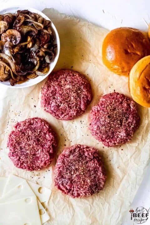 Four ground beef patties, sauteed mushrooms and onions in a white bowl, two brioche buns, and a stack of Swiss cheese on cream-colored parchment paper
