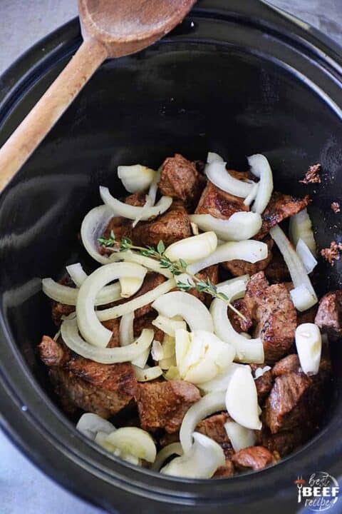 Beef tips in the slow cooker covered by onions and a sprig of fresh thyme