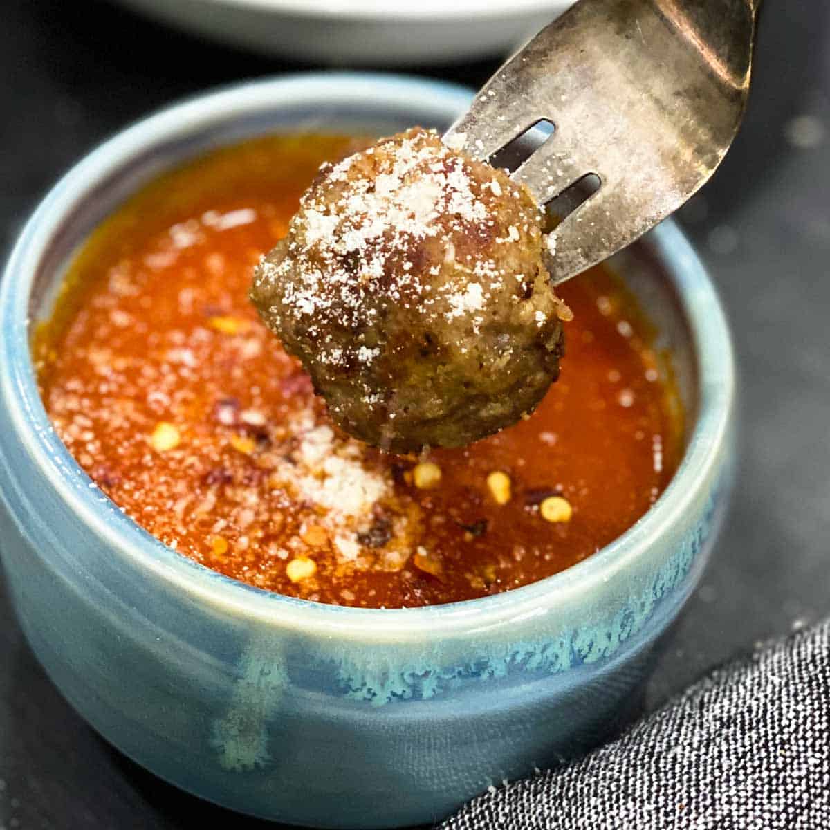 Single meatball on a fork with Parmesan over a bowl of marinara sauce