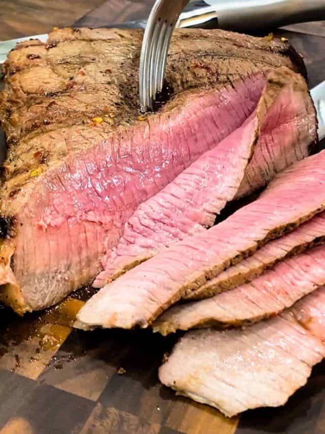 Slicing a grilled london broil on a cutting board with a fork and knife