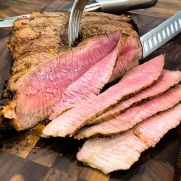 Slicing a grilled london broil on a cutting board with a fork and knife