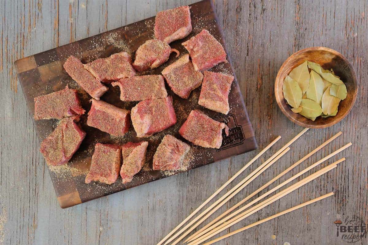 Cubes of seasoned beef on a cutting board with skewers to the side