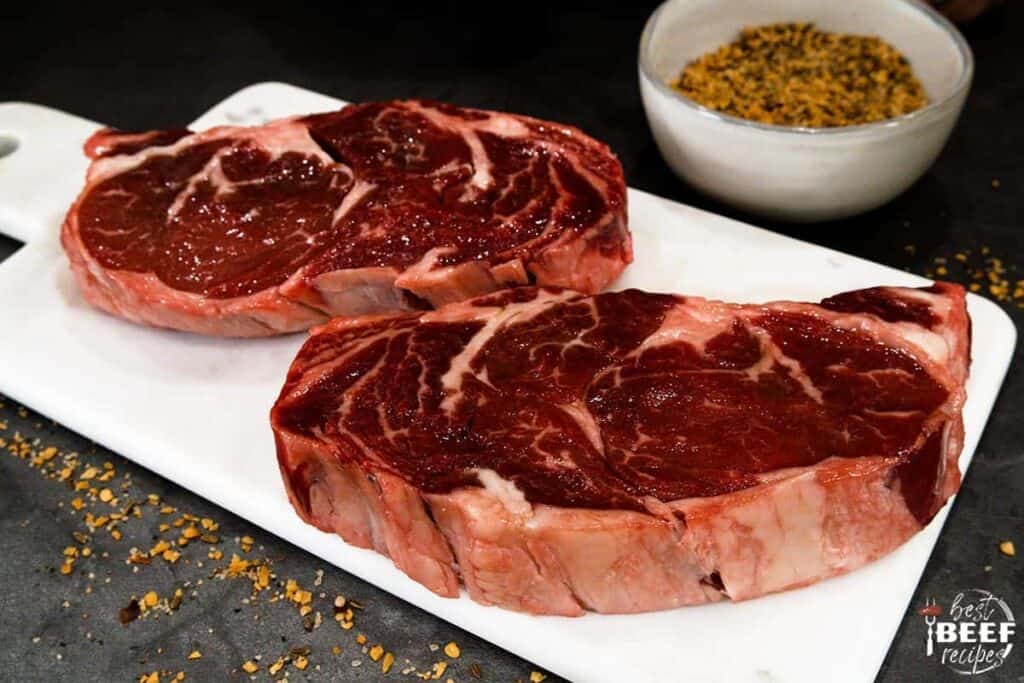 Two rib eye steaks on a white plate with a bowl of seasoning
