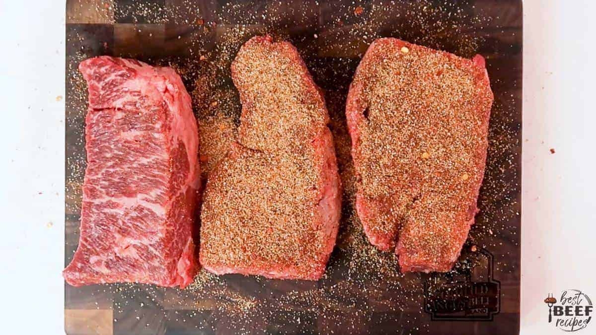 Three steaks on a cutting board with seasoning