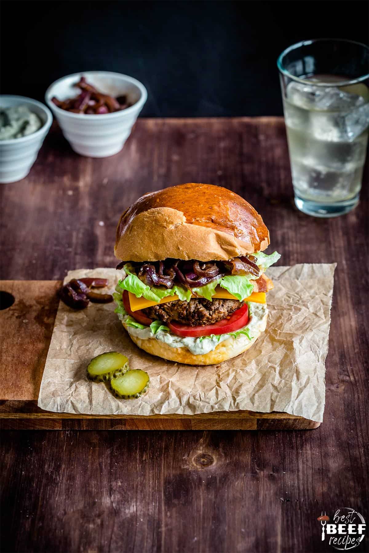 Loaded burger on a sheet of parchment paper with a drink in a glass and toppings in cups