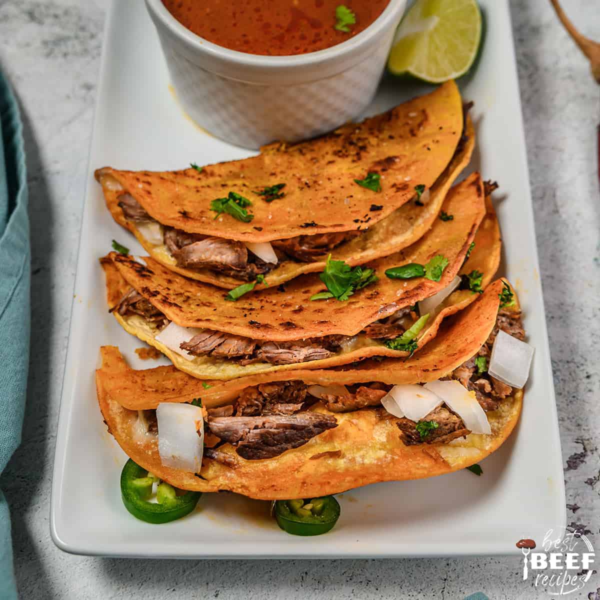 Beef birria tacos on a white platter with birria stew in a bowl