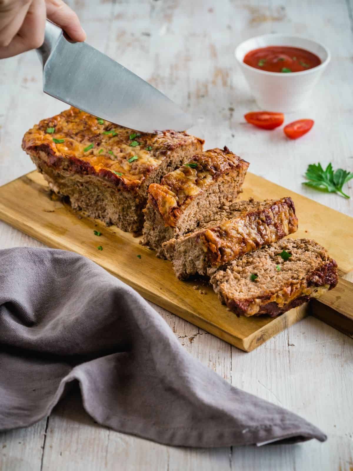 Cheesy meatloaf on a cutting board with 2 slices and being sliced.