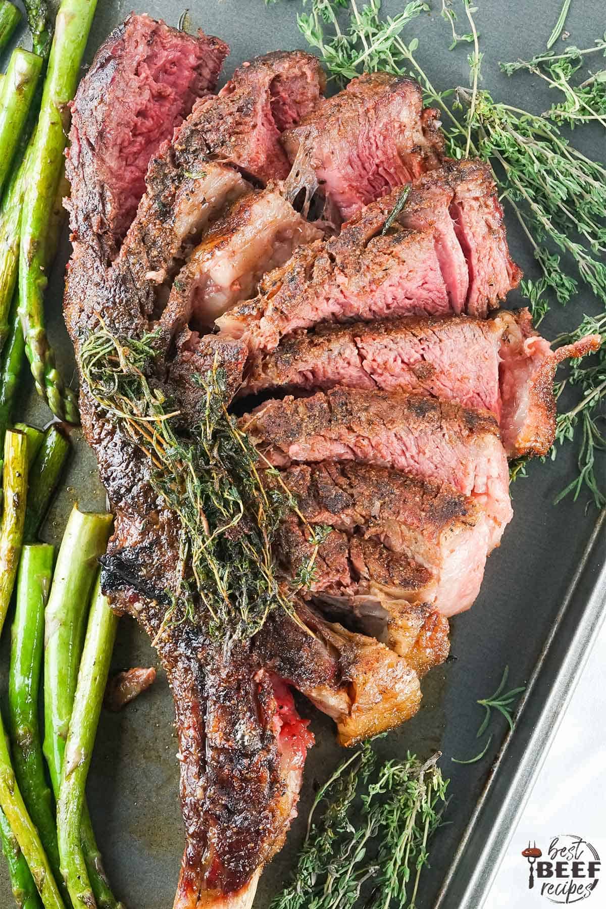 Sliced tomahawk steak overhead shot on a tray with asparagus and thyme