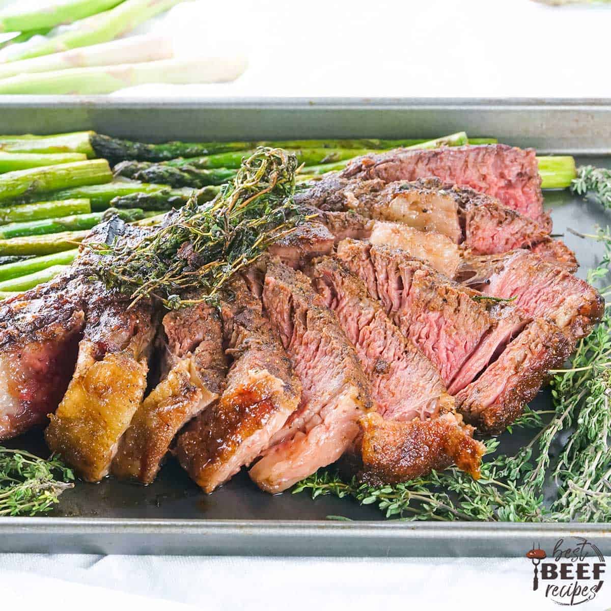 Sliced tomahawk steak on a tray with asparagus and thyme