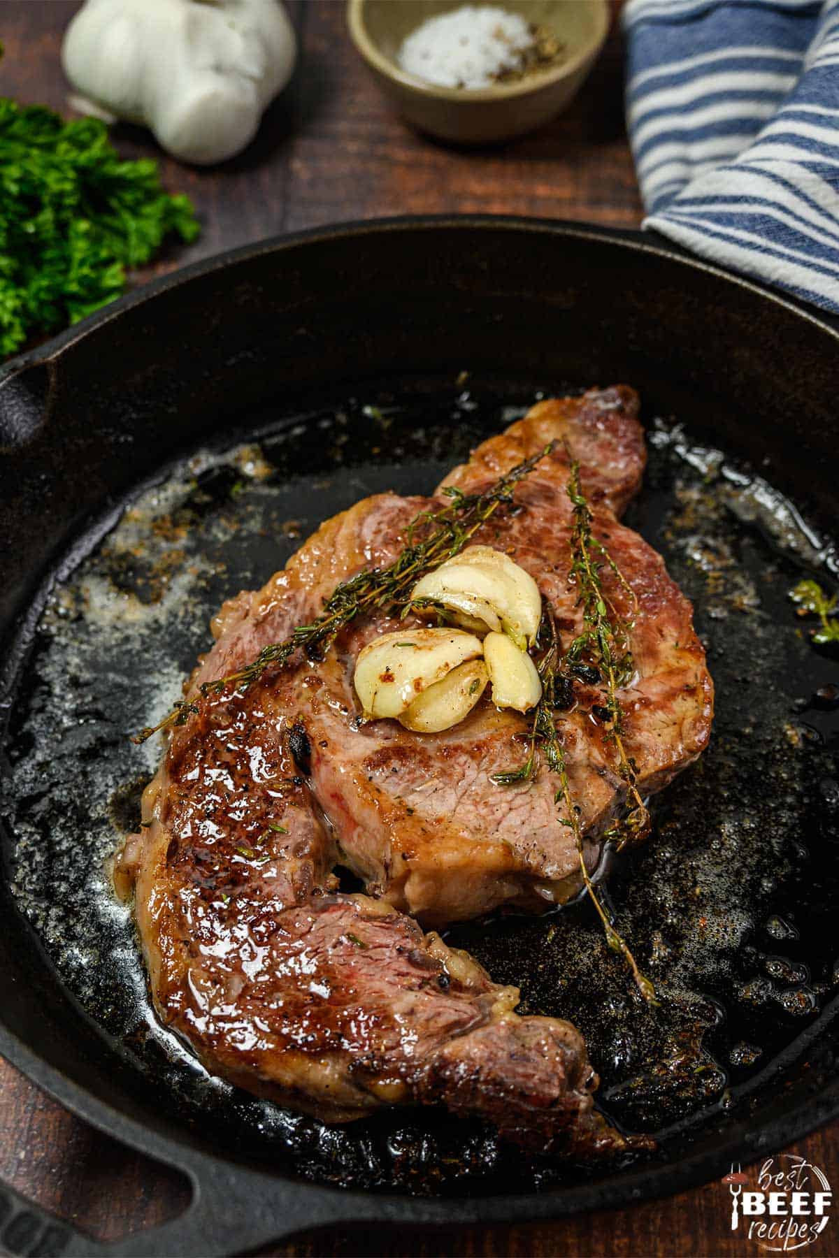 Seared ribeye steak in a pan with cloves of garlic and herbs on top