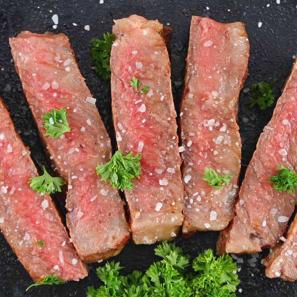 slices of gorgeous medium-rare steak on a black background with coarse salt and parsley