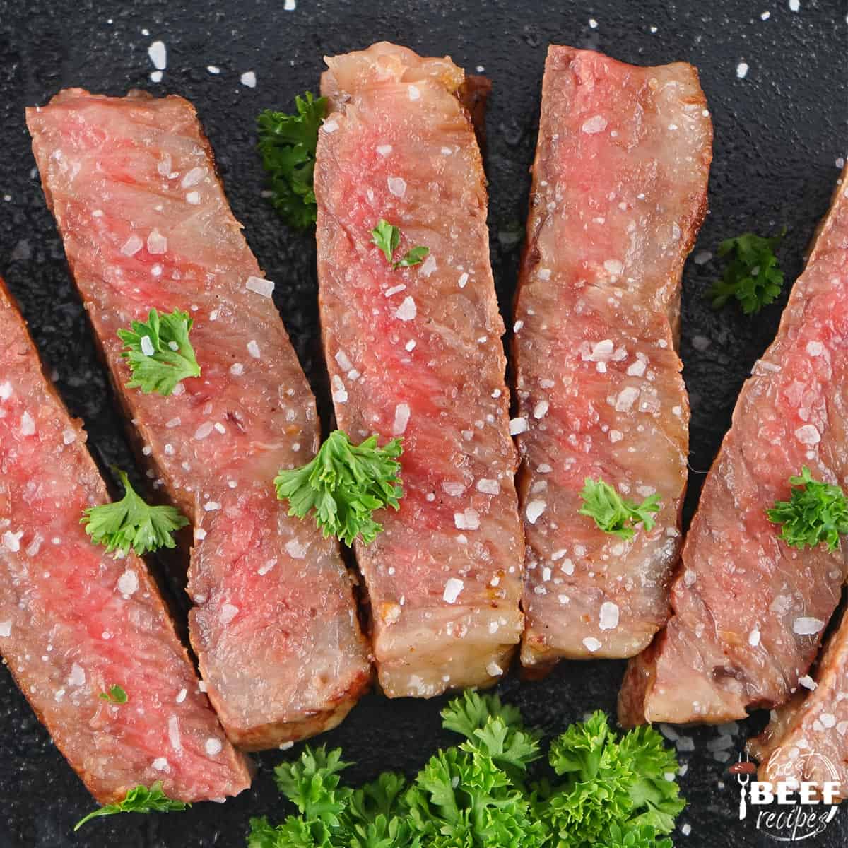 slices of gorgeous medium-rare steak on a black background with coarse salt and parsley