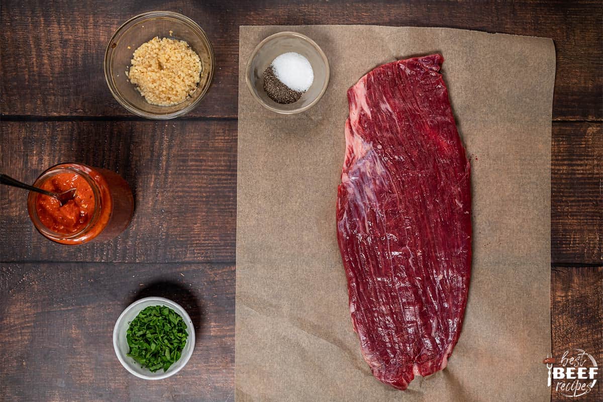 flank steak on a sheet of parchment paper with harissa sauce, garlic, salt and pepper, and parsley in bowls