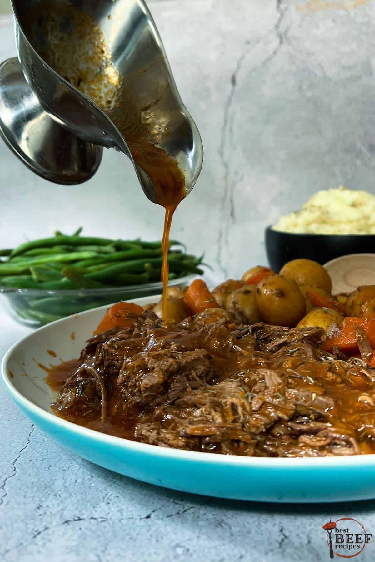 pouring gravy over instant pot pot roast on a plate with carrots and potatoes, in front of a bowl of green beans and mashed potatoes