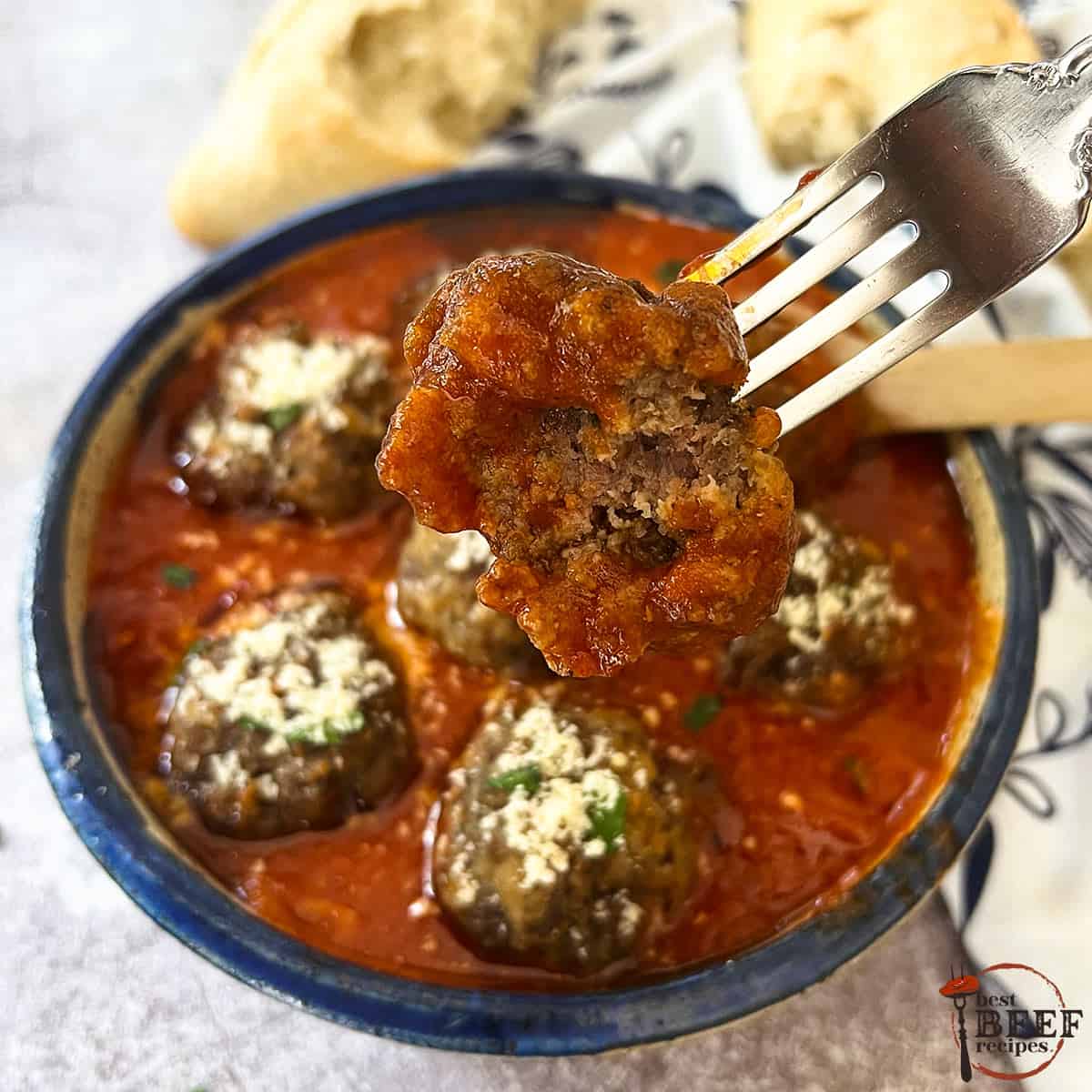 italian meatballs in red sauce in a blue bowl with a metal fork holding half a meatball and bread on the side.