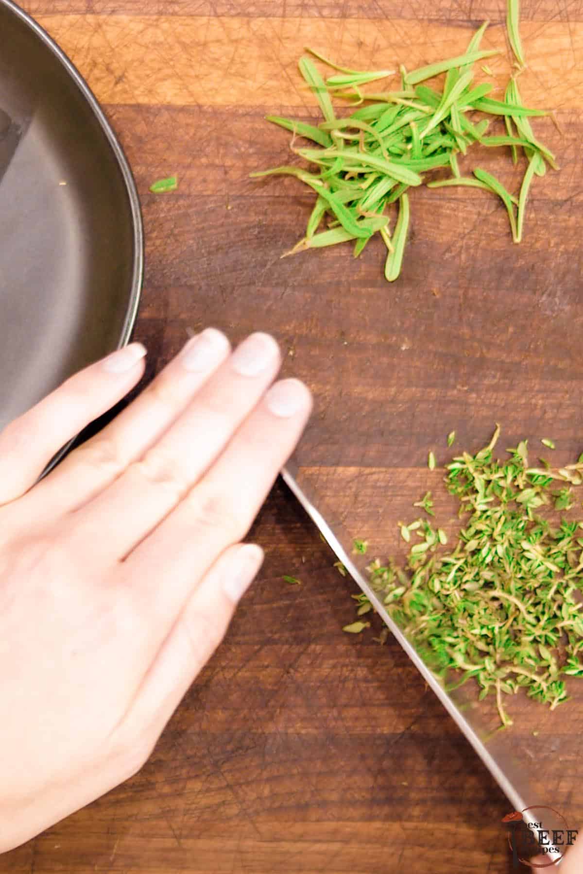 A hand holding a knife to chop thyme on a cutting board