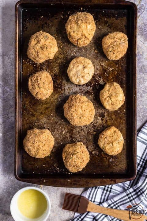a baking sheet full of papas rellenas next to a bowl of melted butter and a butter brush