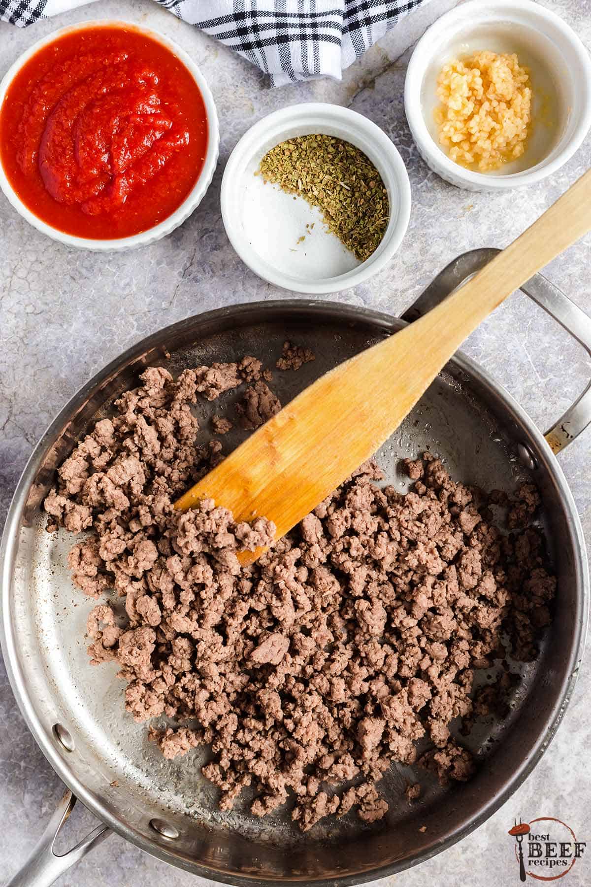 a frying pan with browned ground beef next to separate bowls of tomato sauce, oregano and garlic