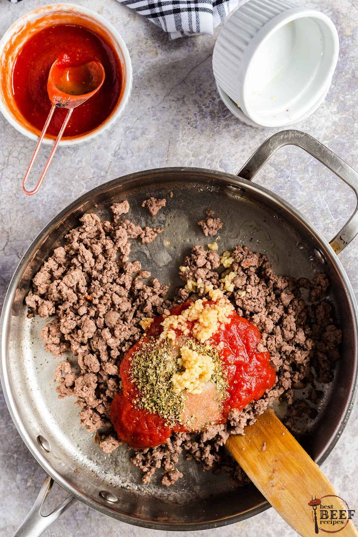 a frying pan with browned ground beef topped with tomato sauce, oregano, salt, and garlic
