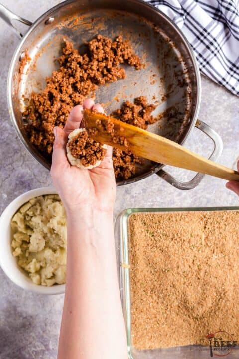 A hand holding a handful of mashed potatoes with ground beef in the center, over dishes of ground beef, mashed potatoes and breadcrumbs