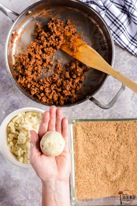 a hand holding a rolled and shaped papas rellenas over separate dishes of mashed potatoes, breadcrumbs and ground beef