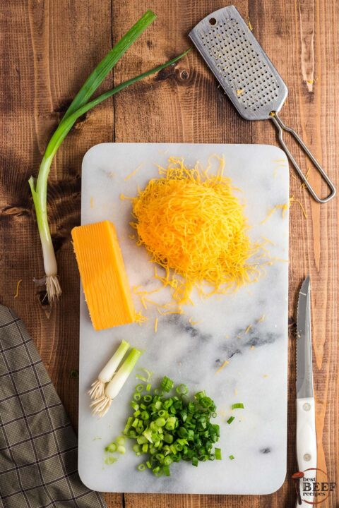 shredded cheese on a cutting board with sliced green onions next to the knife and a grater