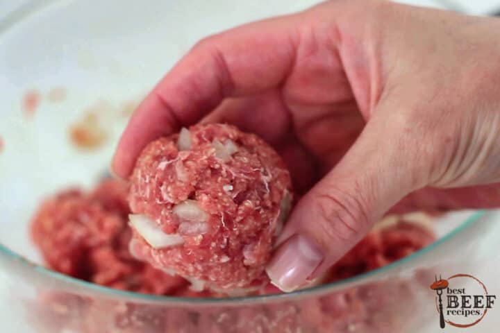 raw meatball mixture being formed into balls by hands over a glass bowl of meatball mix