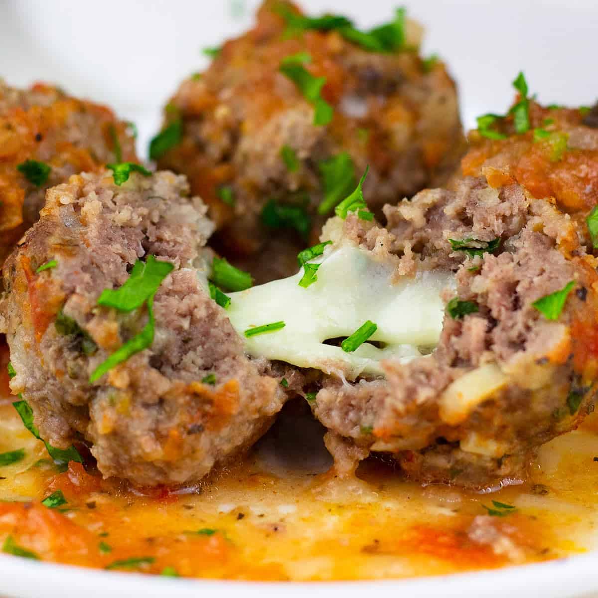 a plate full of meatballs and sauce where the front meatball is split open with mozzarella cheese pulls between the two halves