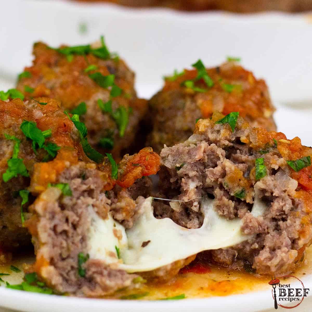 a closeup of a mozzarella stuffed meatball cut in half with mozzarella melted between the two halves, on a plate with more meatballs