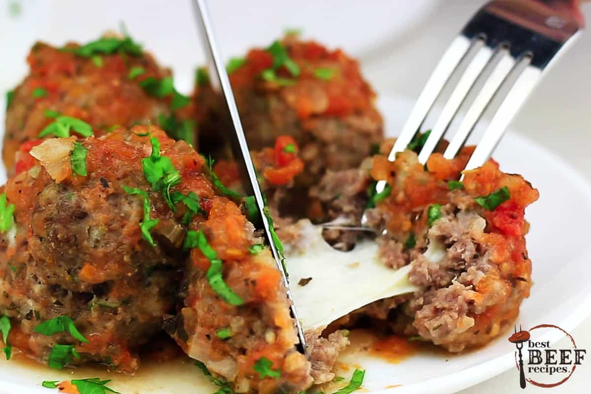 a mozzarella stuffed meatball being cut open by a fork and knife on a plate of meatballs