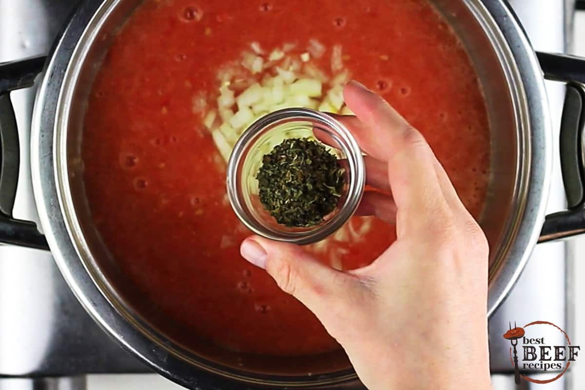 a pot full of tomato sauce with a hand holding a glass bowl of seasoning over the pot