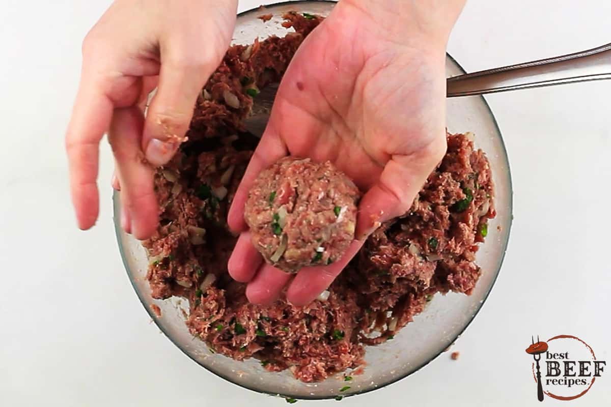 hands holding a rolled meatball over a dish of meatball mixture on a white background