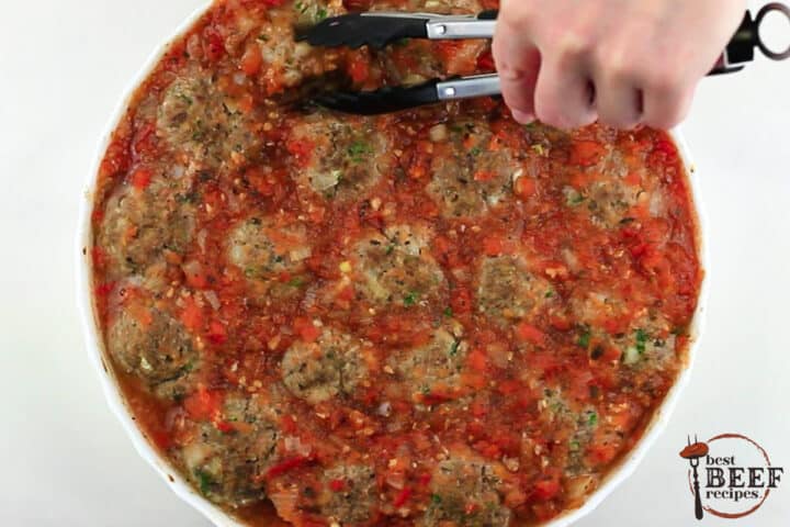 A white dish of fully cooked mozzarella stuffed meatballs with tongs reaching for a meatball