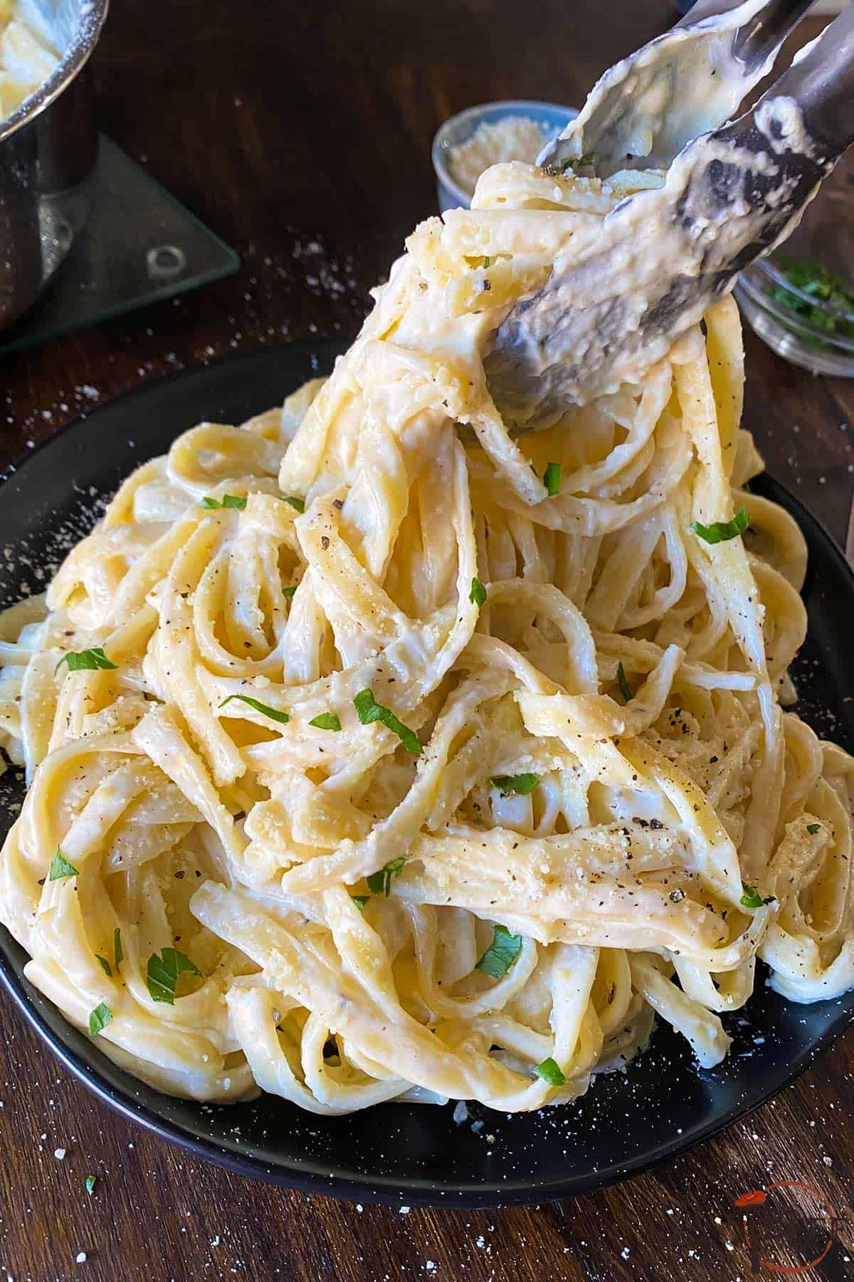 tongs placing fetttuccine noodles on to a plate with parlsey garnish