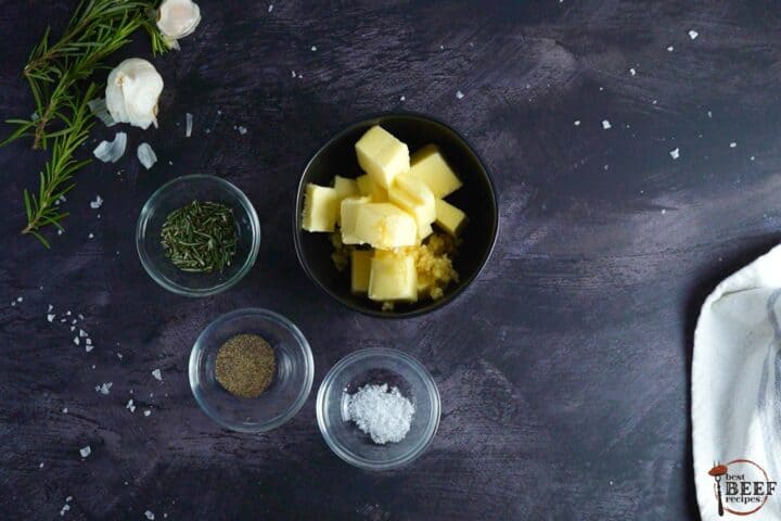 butter and garlic in a black bowl next to three bowls of rosemary, salt, and pepper