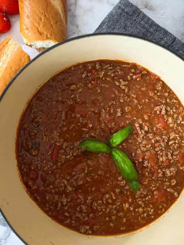a big pot of sugo di carne with basil leaf garnish next to bread, tomatoes, and extra parmesan cheese