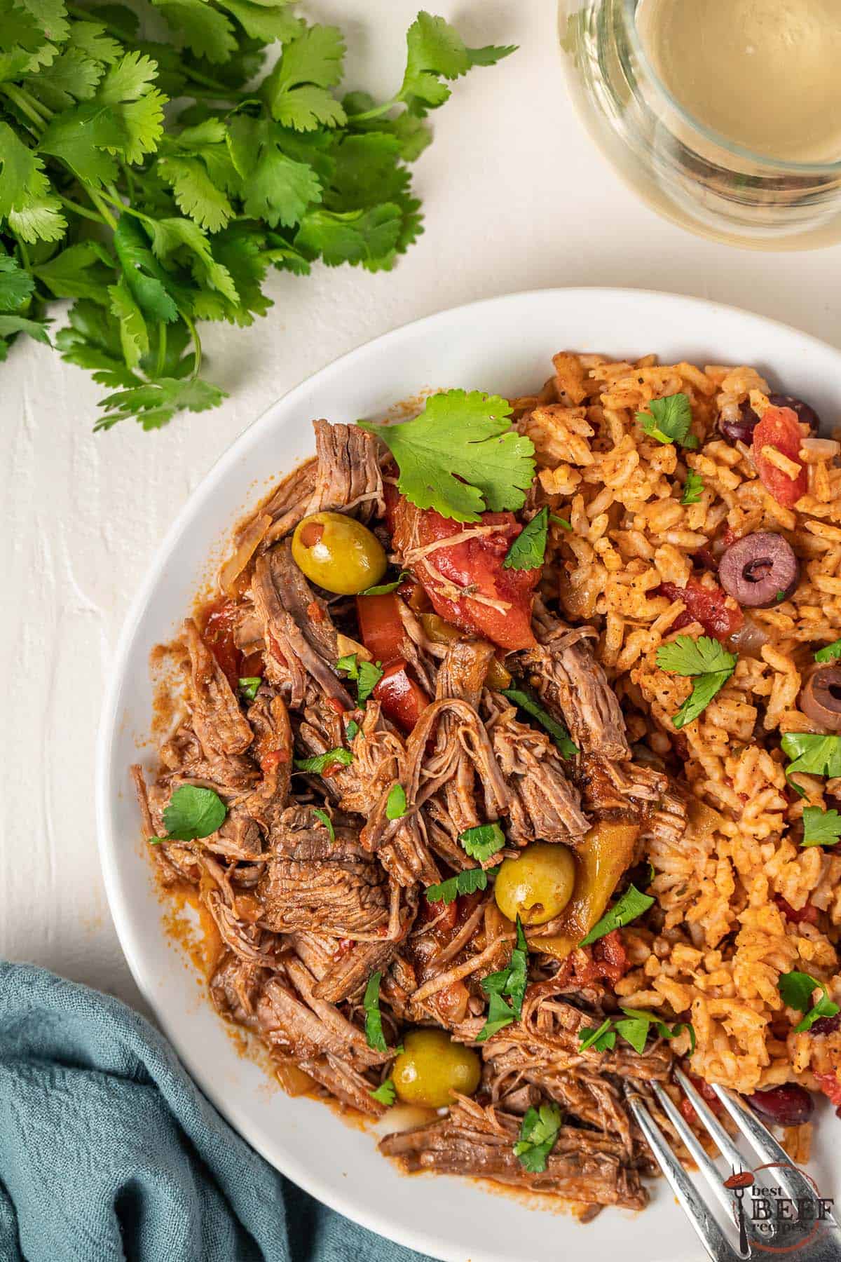 ropa vieja and beans and rice on a plate next to white wine and a bunch of cilantro