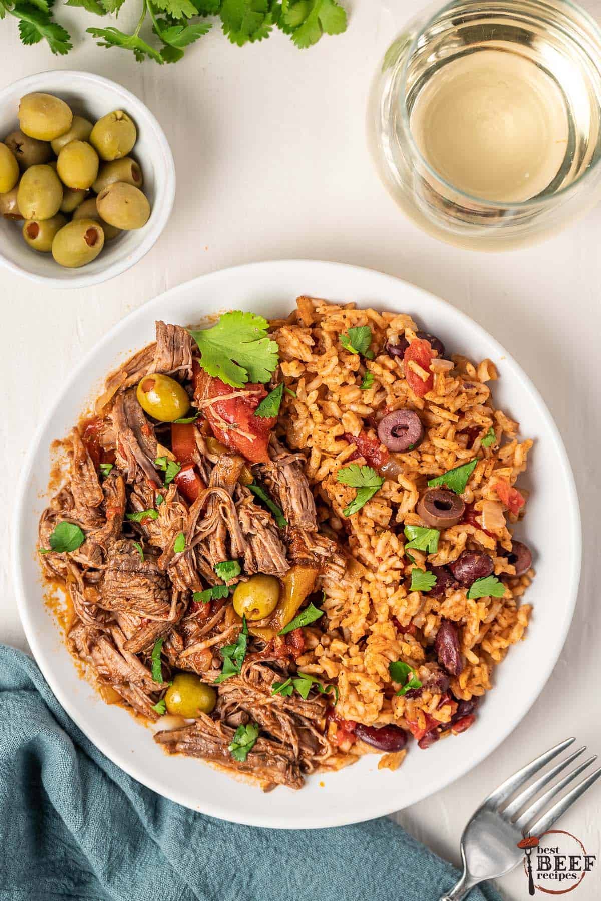 a dinner plate of ropa vieja and beans and rice with a cup of wine and a dish of olives