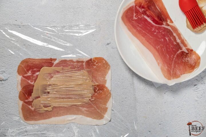 prosciutto laid out flat on plastic wrap with dijon mustard slathered on