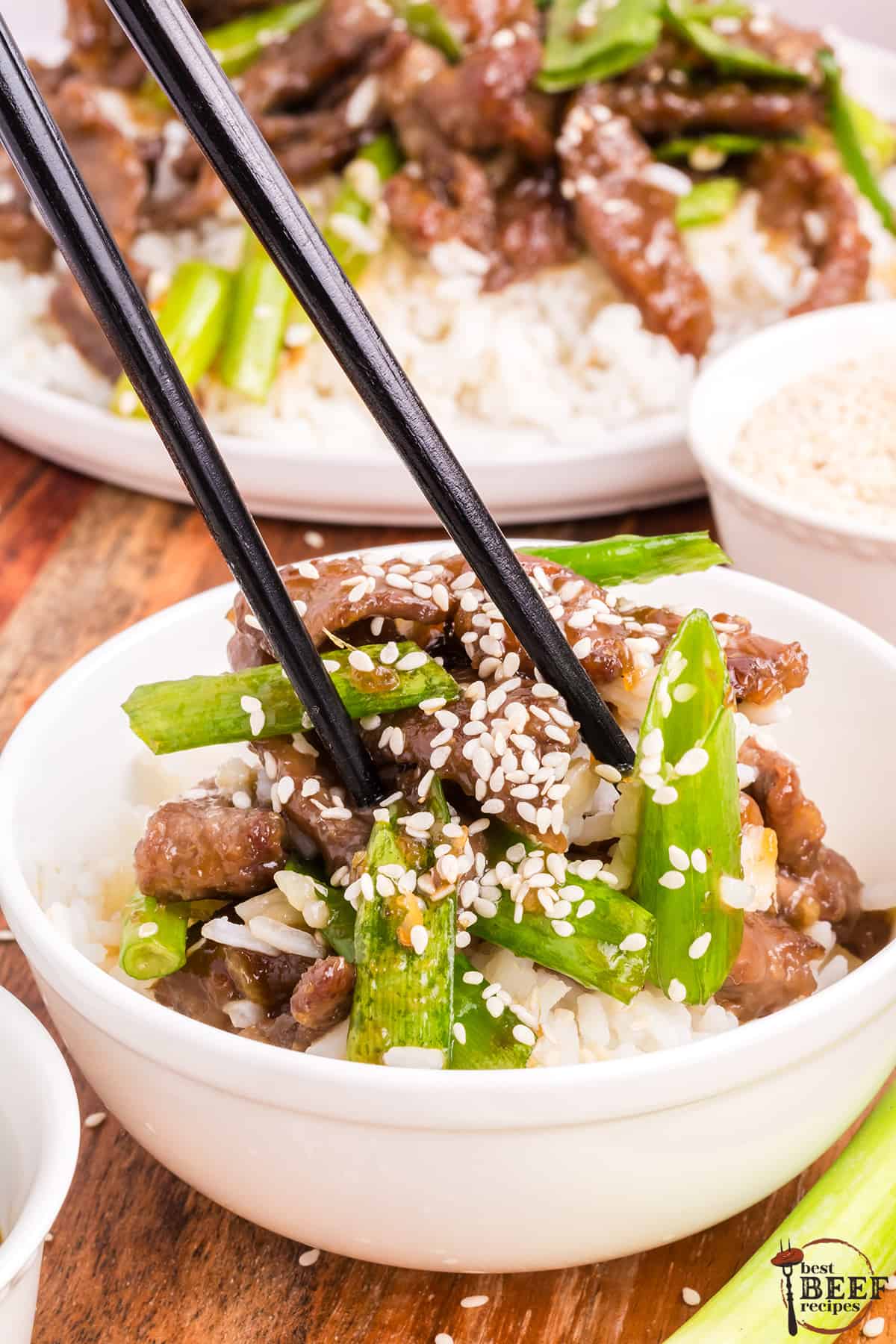 mongolian beef in a bowl with green onions, sesame seeds, and rice; chopsticks lifting a piece