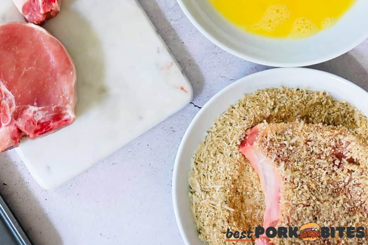 a pork chop being dipped in coating next to a bowl of egg and a cutting board with raw pork chops