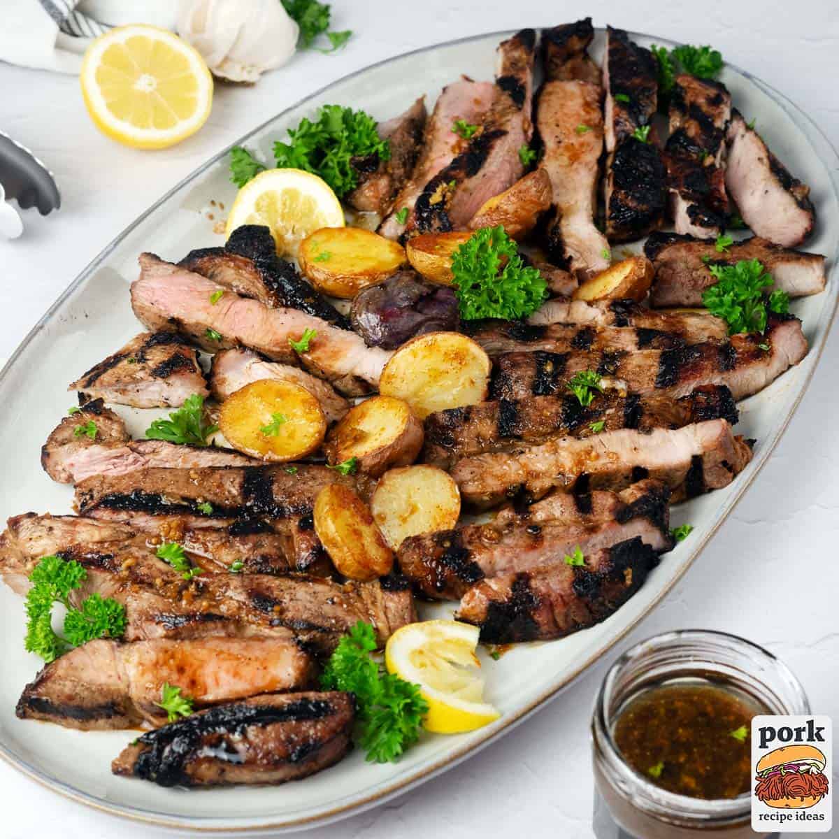 grilled pork steak sliced on a plate with potatoes, herbs and lemon wedges